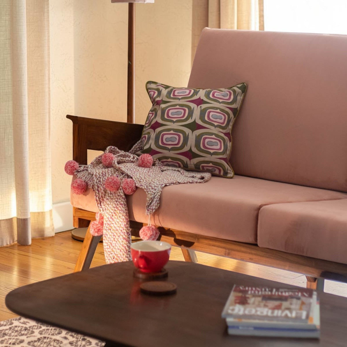 Homecrest Culworth mid-century slim teak wood sofa in soft pink upholstery with a patterned cushion and pom-pom throw.