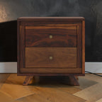 Homecrest Piera mid-century bedside table in dark sheesham wood with two drawers, brass knobs, and tapered legs.