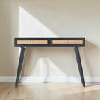 Homecrest Isla console table in black with two woven rattan cane drawers and gold handles against a beige wall.
