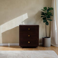 Homecrest Mersham bedside table in dark solid hardwood with three drawers and gold knobs in a sunlit room.