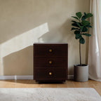 Homecrest Mersham bedside table in dark solid hardwood with three drawers and gold knobs in a sunlit room.