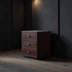 Homecrest Mersham bedside table in dark solid hardwood with three drawers and gold knobs in a moody bedroom setting.