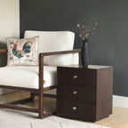Homecrest Mersham bedside table in dark solid hardwood with three drawers next to a modern cream armchair