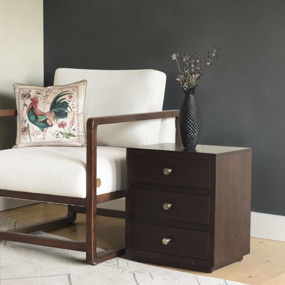 Homecrest Mersham bedside table in dark solid hardwood with three drawers next to a modern cream armchair